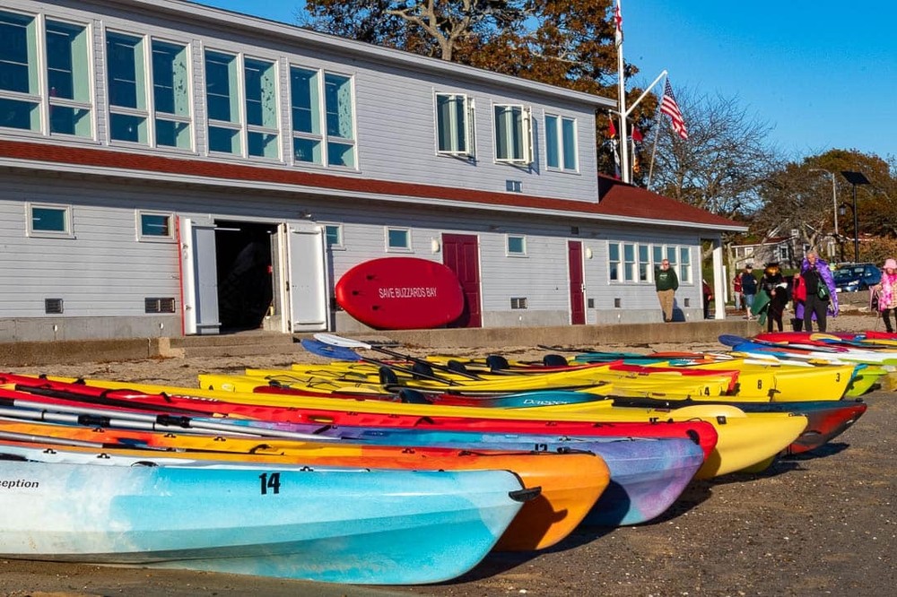 Colorful kayaks lined up on the beach in front of the Onset Bay Center, ready for participants at the Halloween Paddle Parade event.